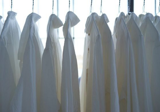 A row of white hospital gowns hanging on a garment rack, backlit by a window.